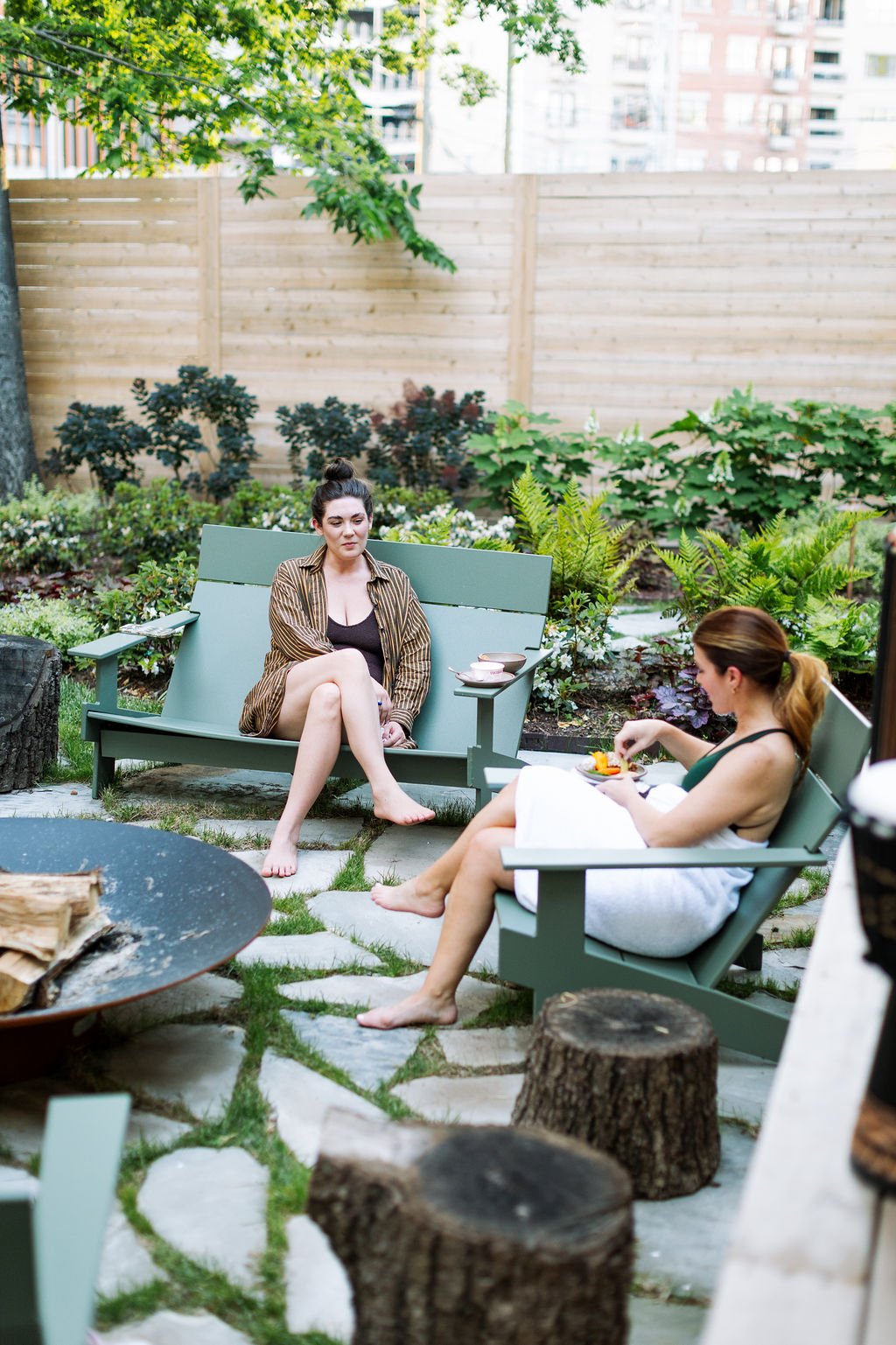Two women relaxing outdoors in a garden, one sitting on a green bench with a towel wrapped around her, and the other in a chair with a towel, eating a meal. The garden has lush plants and a wooden fence, with buildings visible in the background.