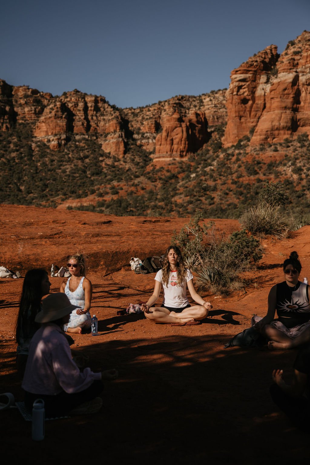 Group of women practicing meditation outdoors on red desert ground with red rock formations and cliffs in the background, some sitting in lotus position and others relaxing.