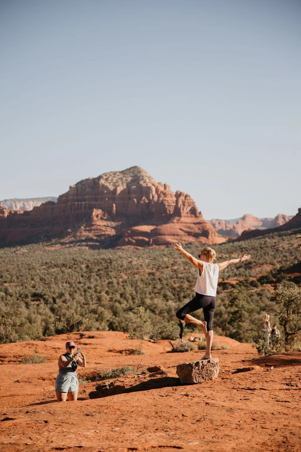 A woman balancing on a rock with arms outstretched in a desert landscape with red rocks and mountains, two other women taking photos in the background.