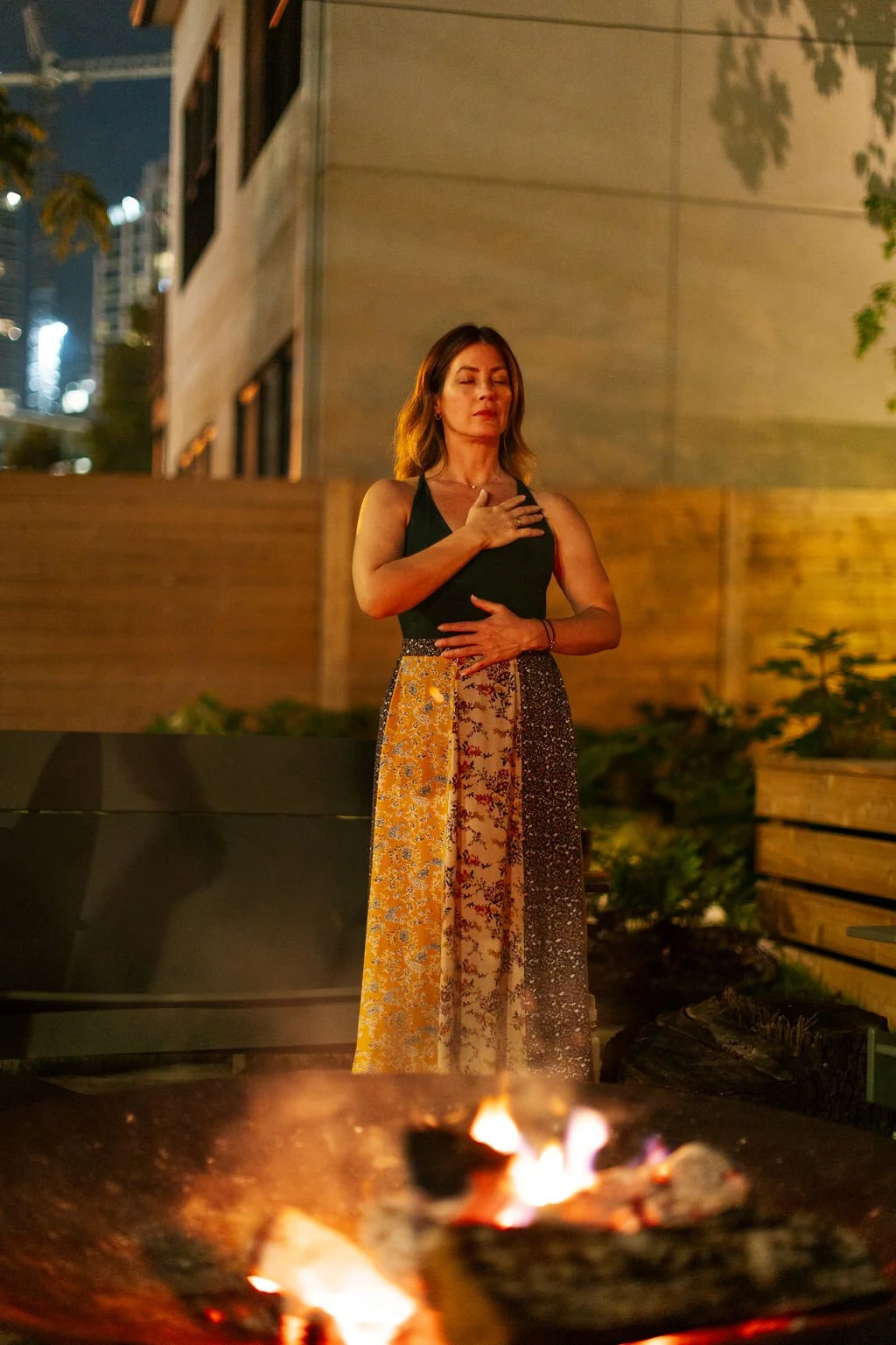 A woman standing outdoors at night with her eyes closed, one hand on her chest, and the other on her stomach, near a fire pit.
