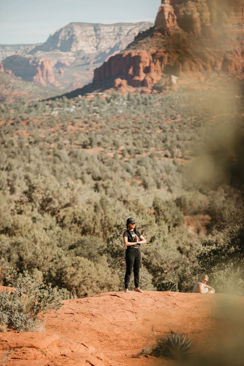 Woman standing on a red rock formation in a desert landscape with red mountains and sparse vegetation in the background.