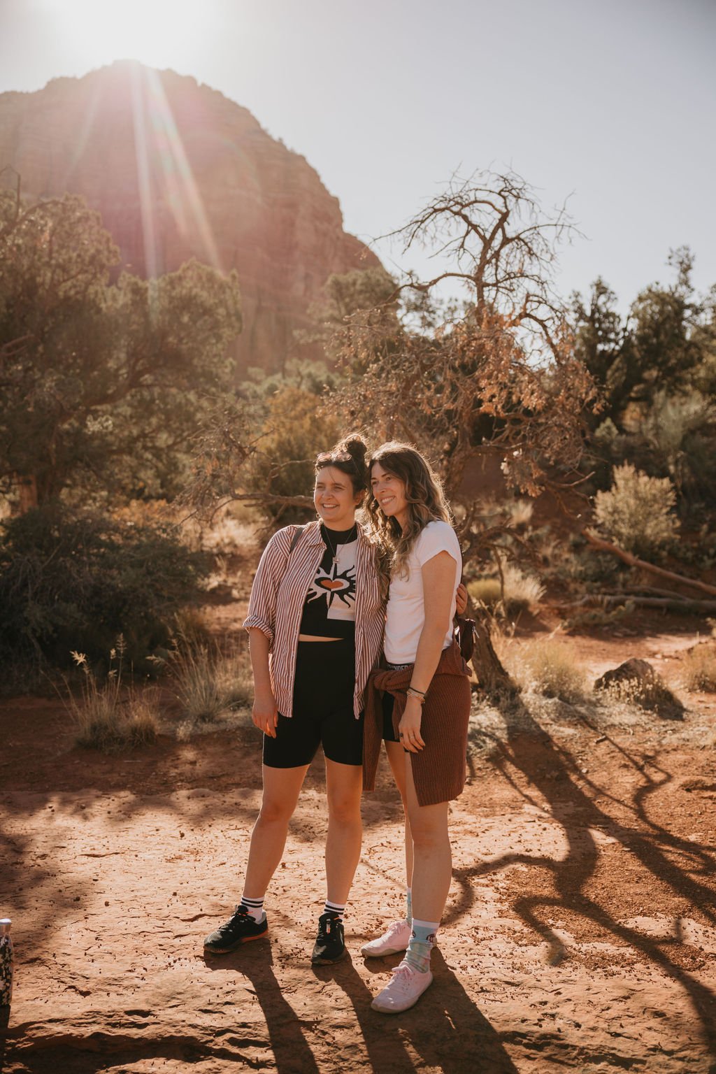 Two women standing close together outdoors with desert landscape, trees, and mountain in the background during sunset, smiling.