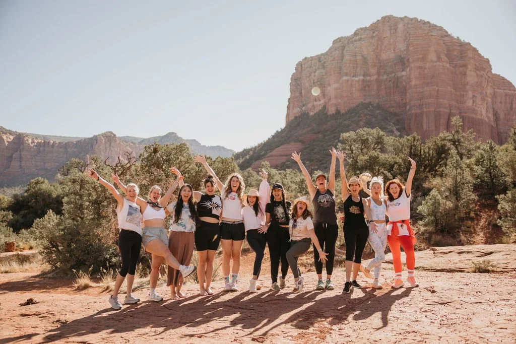 Group of women cheering and smiling in the desert with red rocky cliffs and mountains in the background.