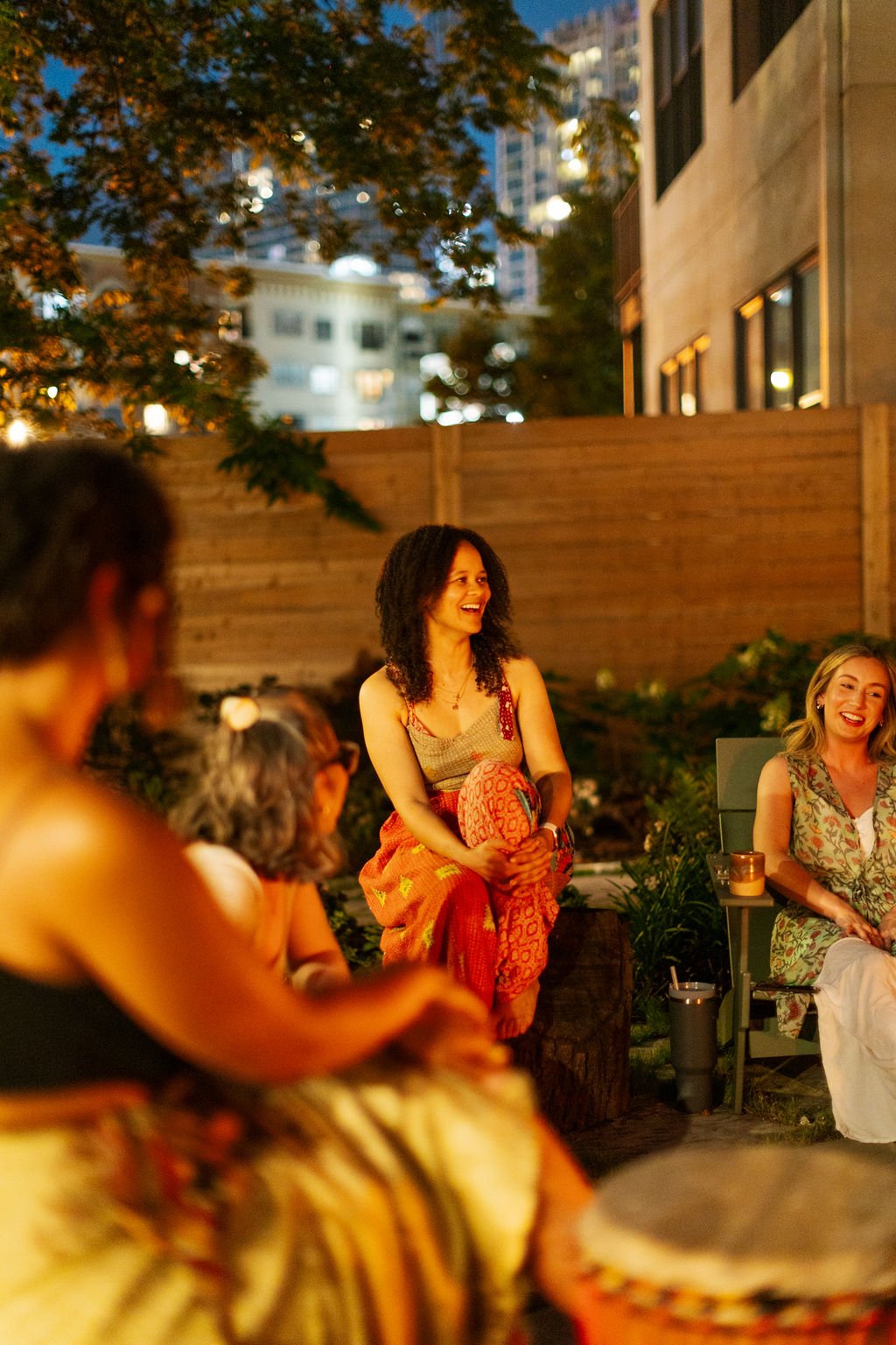 Group of women enjoying an evening outdoor gathering, smiling and sitting around a firepit with city buildings and trees in the background.