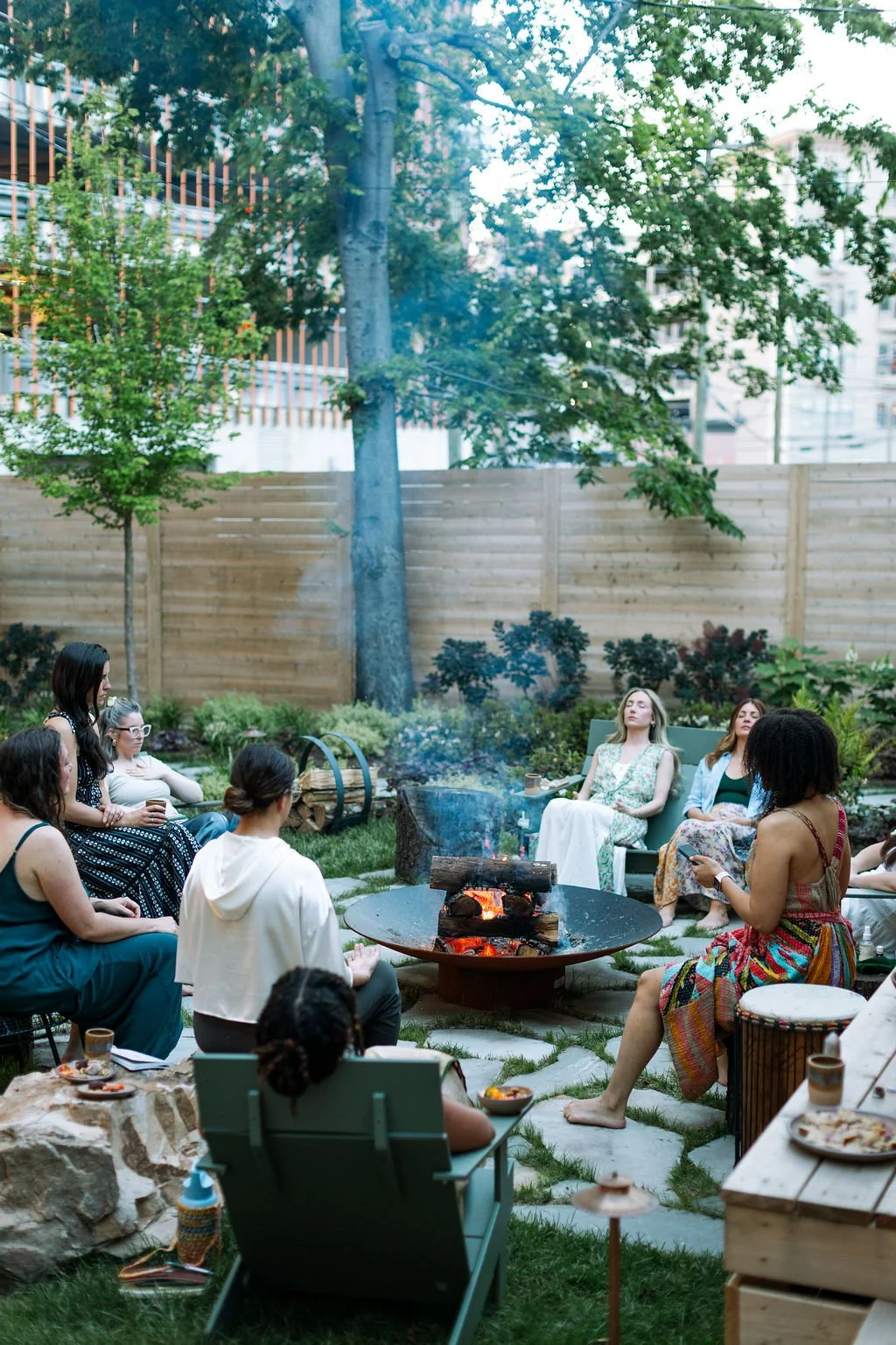 A group of women gathered around a fire pit in a backyard garden, relaxing and socializing during an outdoor gathering or barbecue.