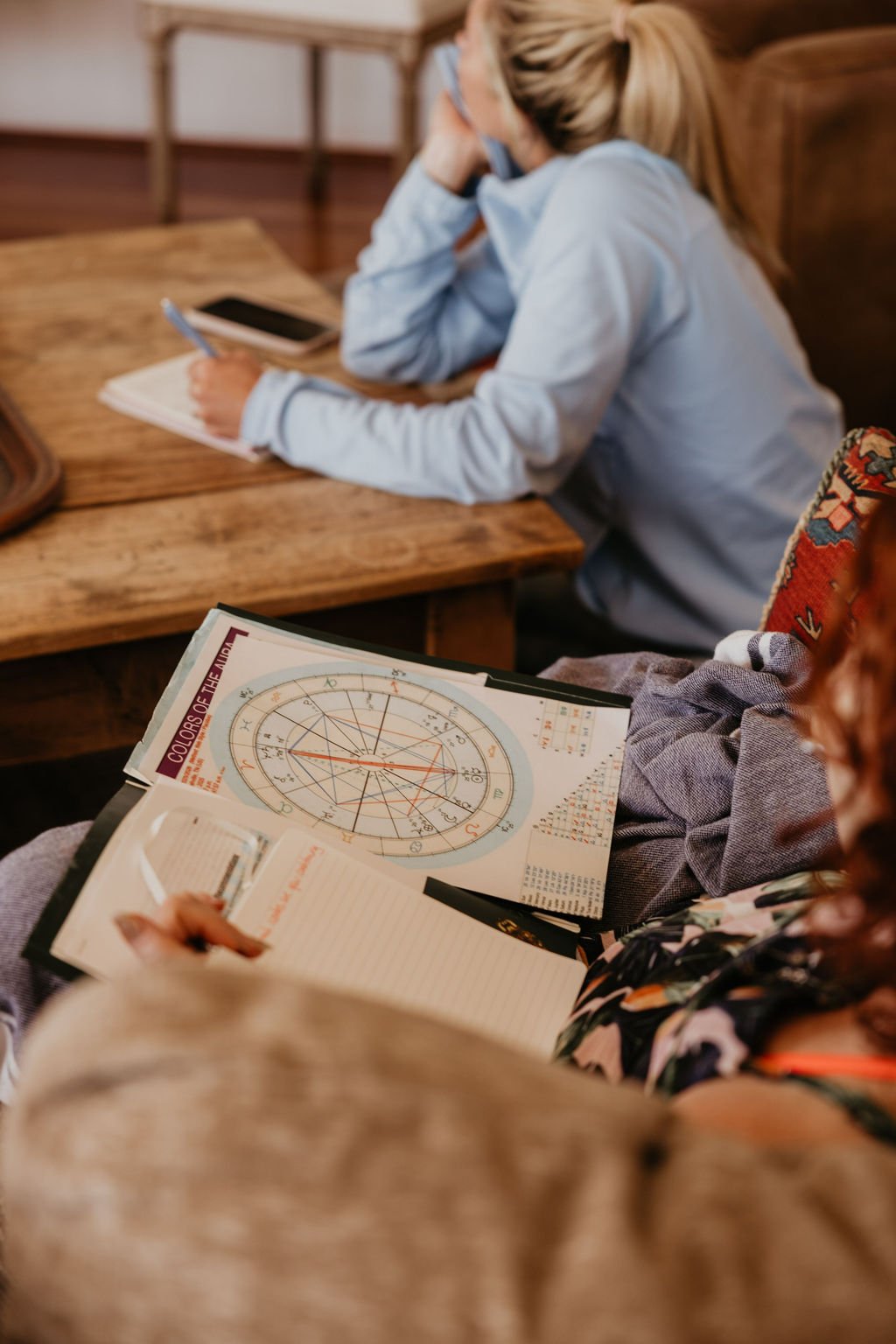 Person holding a book titled 'Colors of the Moon' and a notebook, with an astrological chart visible on the book. In the background, a woman wearing a light blue hoodie and glasses is writing in a notebook at a wooden table.