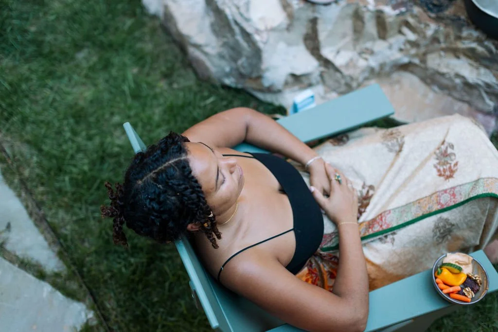 A woman with braided hair and tattoos relaxing outdoors on a blue lawn chair, covered with a floral-patterned blanket, next to a stone wall and a small table with a plate of vegetables.