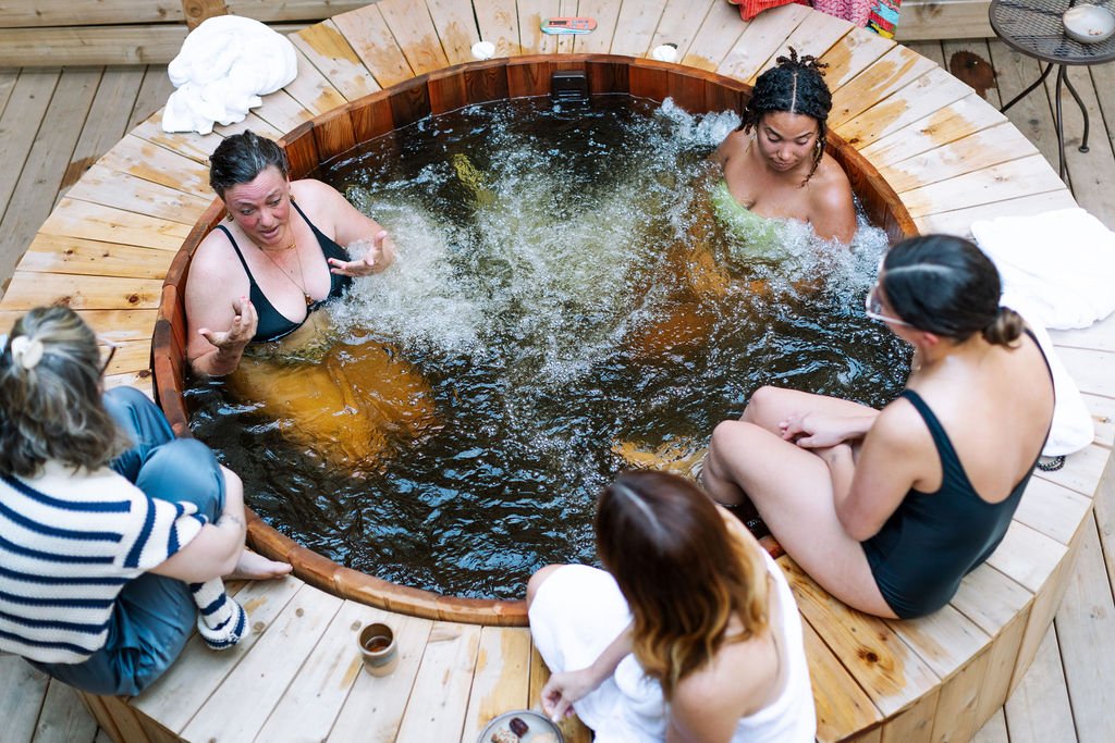Group of women sitting in a wooden hot tub outdoors, engaging in conversation and relaxation.
