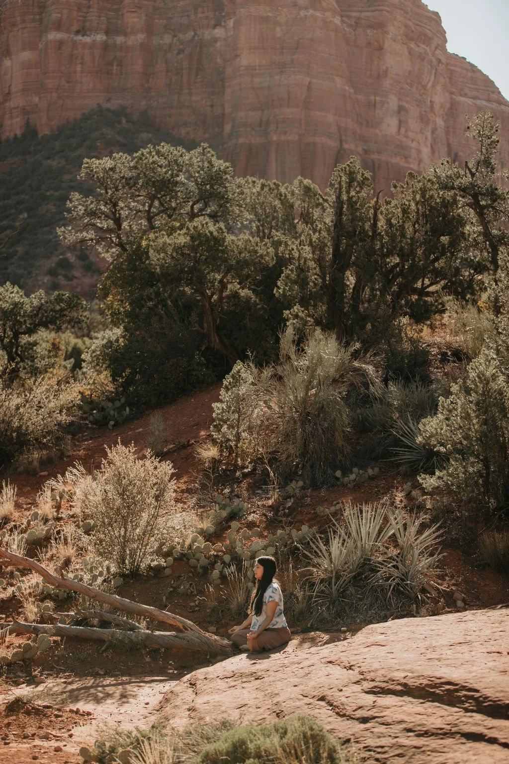 A woman meditating outdoors in a desert landscape with bushes, cacti, and a large rocky cliff in the background during daytime.