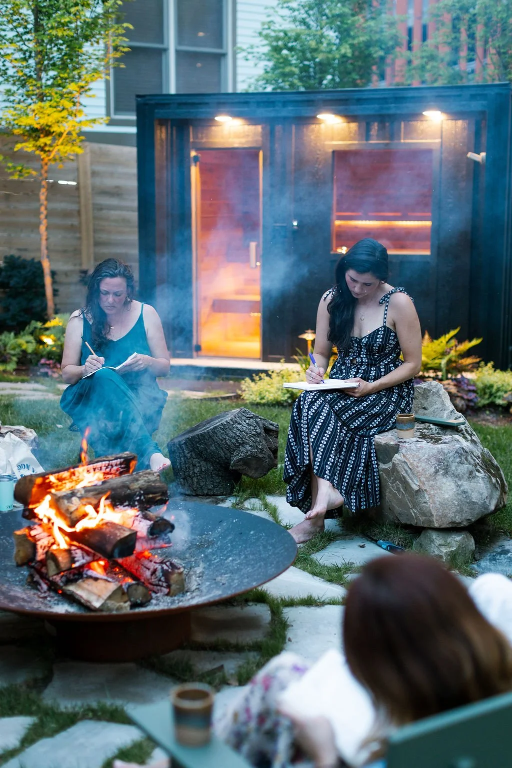 Three women sitting outdoors beside a firepit, writing or drawing in notebooks, with a small black shed and a house in the background during dusk.