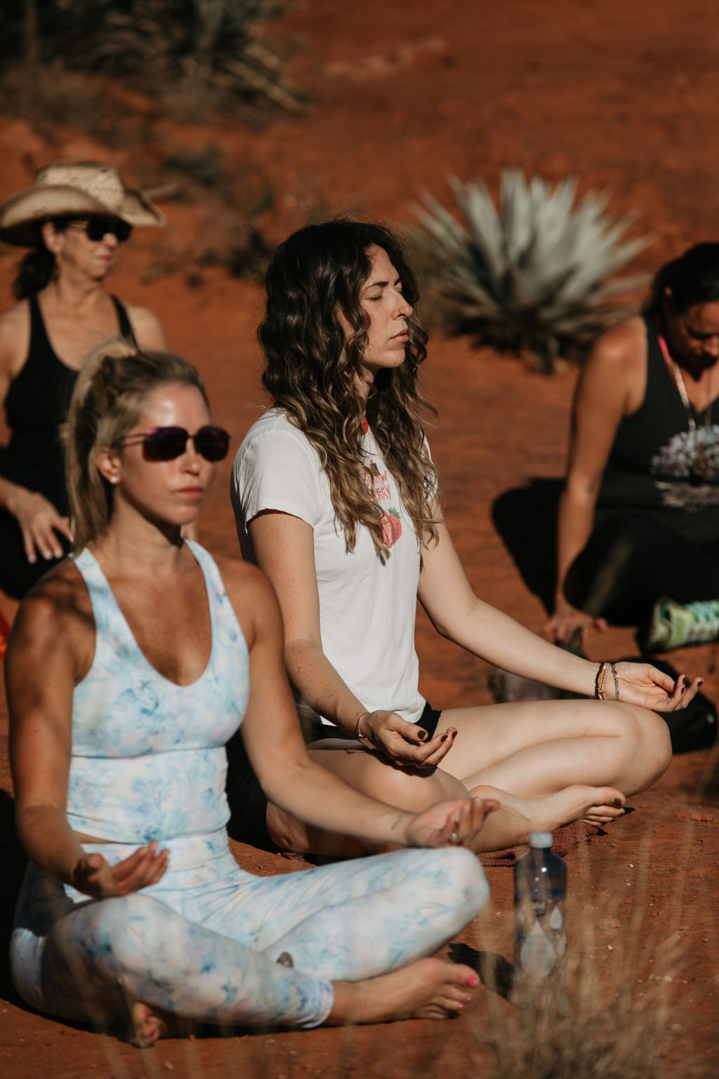 Four women are sitting cross-legged on the ground meditating outdoors in a desert-like environment with sparse vegetation.