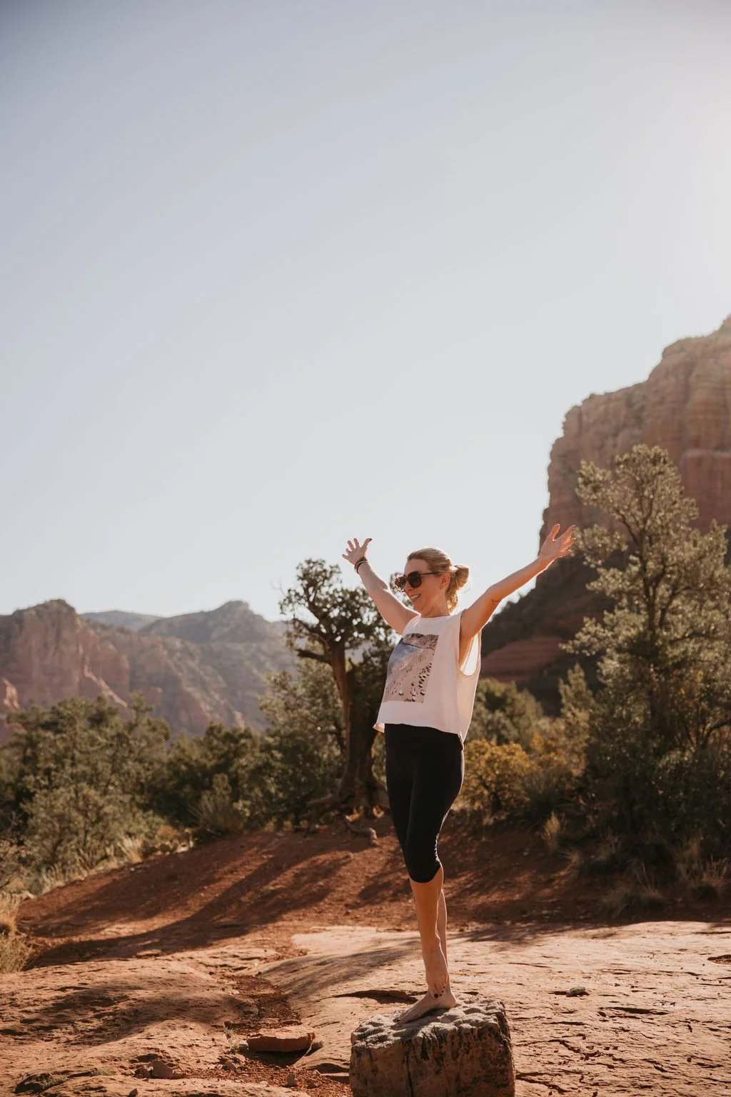 A woman standing barefoot on a rock in a desert landscape with mountains and trees, raising her arms joyfully and wearing sunglasses.