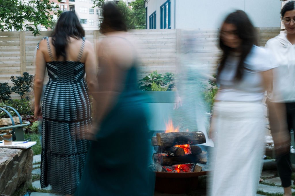 People gathered around a small outdoor fire pit in a backyard with wooden fencing and greenery, some are wearing summer dresses.