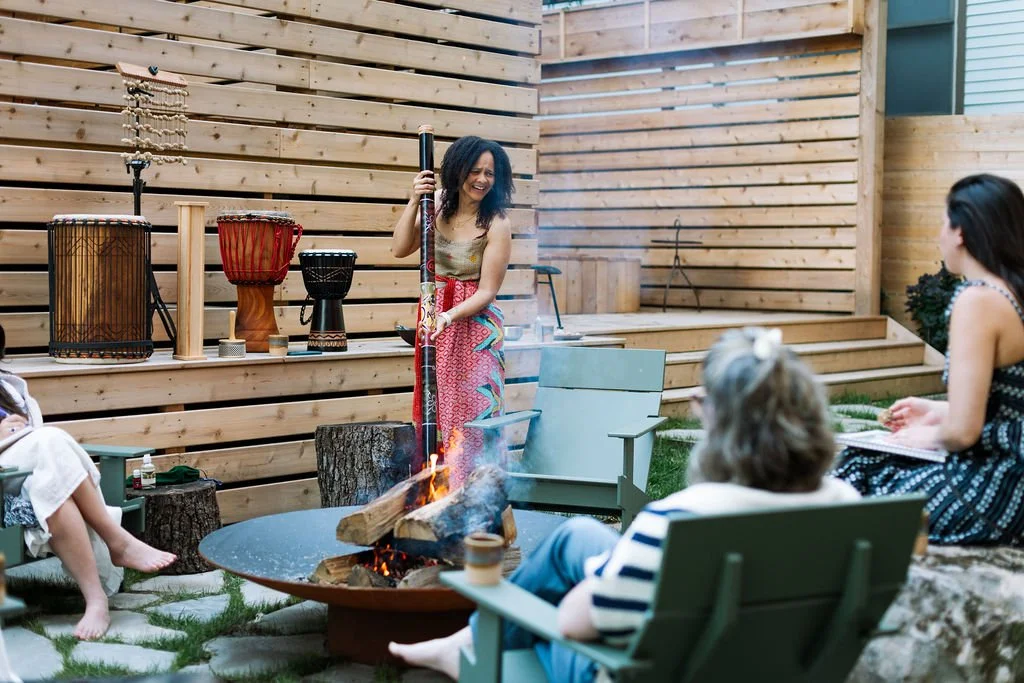 A woman standing by a fire pit, holding a flaming torch, surrounded by three seated women. The scene takes place outdoors with a wooden fence background and various drums displayed on a wooden surface behind the woman.