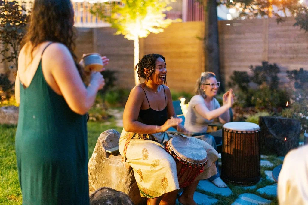 Three women enjoying a backyard gathering, with one woman playing a drum and smiling, two others clapping and one holding a drink, during evening time.