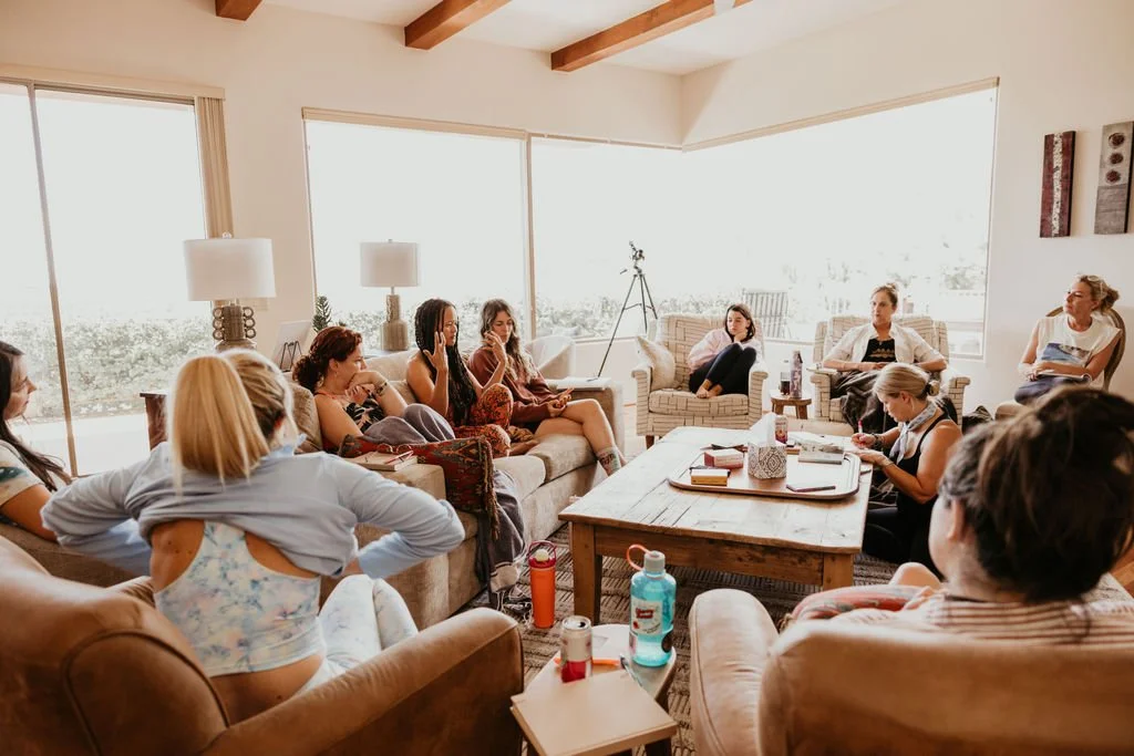 A group of women sitting in a bright living room with large windows, engaging in a discussion or meeting, with some taking notes and others listening attentively.