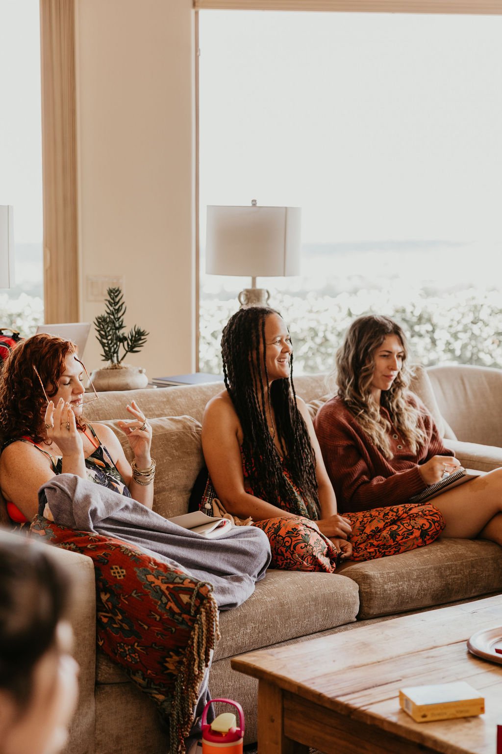 Three women sitting on a beige couch in a brightly lit living room, smiling and engaged in conversation, with a large window revealing a snowy landscape outside.