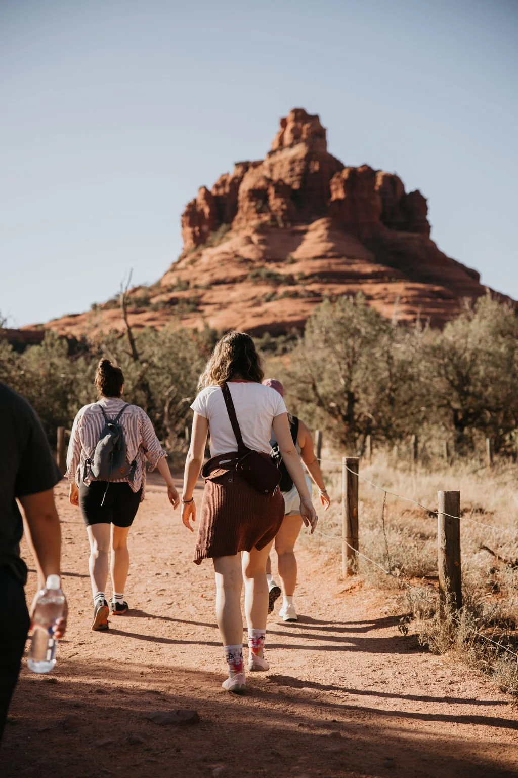 Group of people hiking on a dirt trail in a desert landscape with red rock formations and sparse trees.