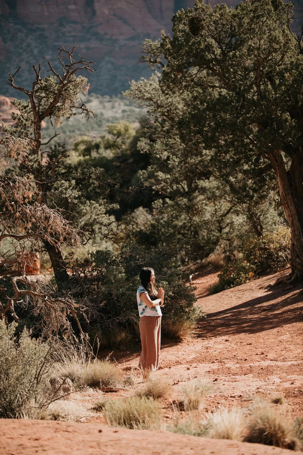 A woman practicing yoga or meditation outdoors in a desert landscape surrounded by trees and rocky terrain.