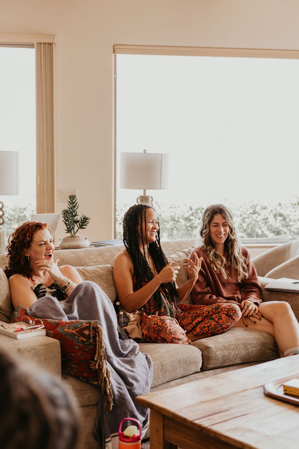 Three women sitting on a beige couch in a living room, smiling and laughing. The woman on the left has red hair and is wrapped in a blanket, the middle woman has dark braids and is wearing a patterned outfit, and the woman on the right has long, wavy