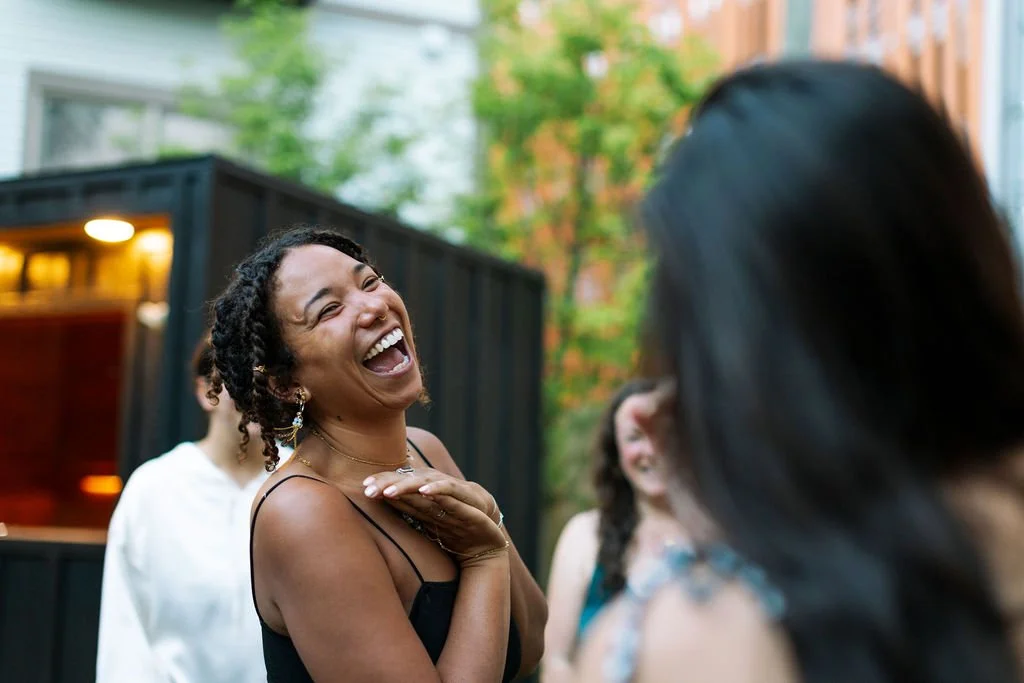 A woman with short curly hair laughing and smiling, wearing a black spaghetti strap top, in an outdoor setting with trees and other people in the background.