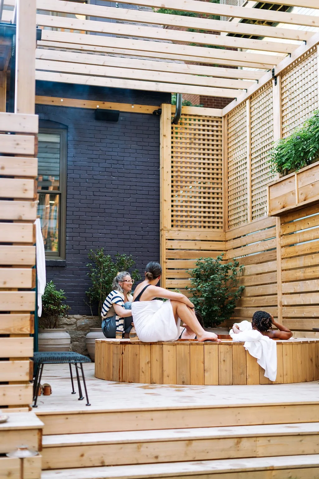 Three women relaxing in a backyard hot tub with wooden walls and plants, one wrapped in a white towel and the other two in towels, during the daytime.