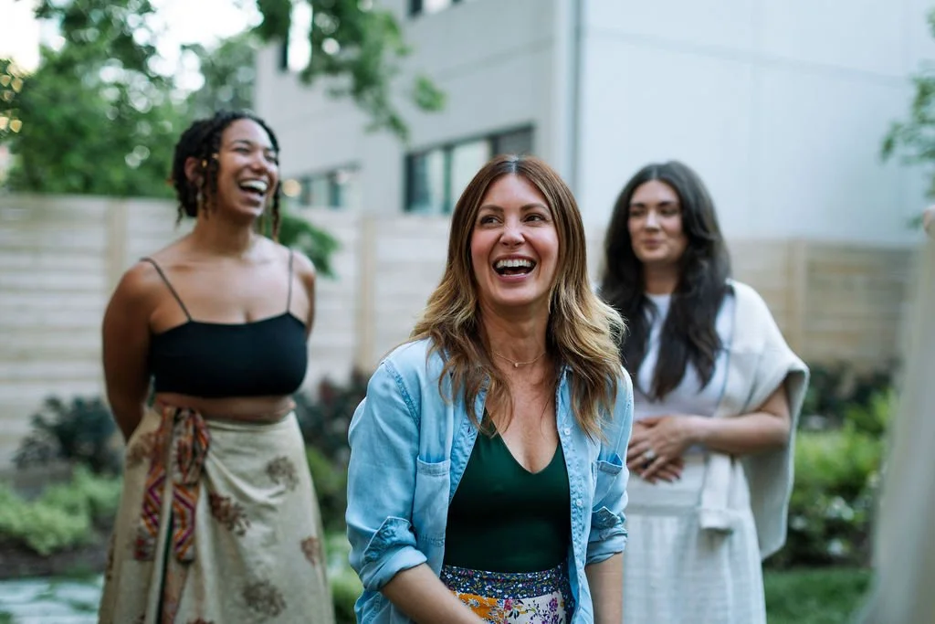 Four women smiling and laughing outdoors in a garden area with trees and a wooden fence in the background.
