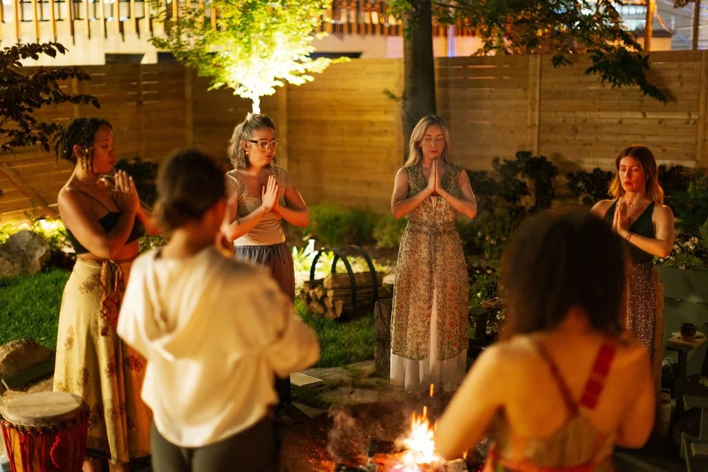 Group of women gathered around a fire pit in a backyard at night, meditating with their hands in a prayer position.