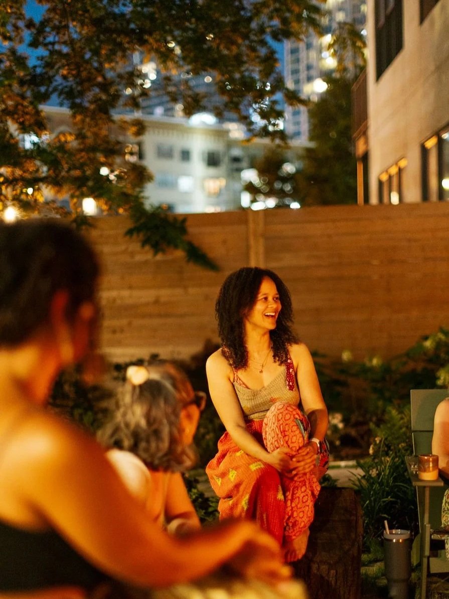 A woman with curly hair sitting outdoors at night, smiling and engaging with a group of people.