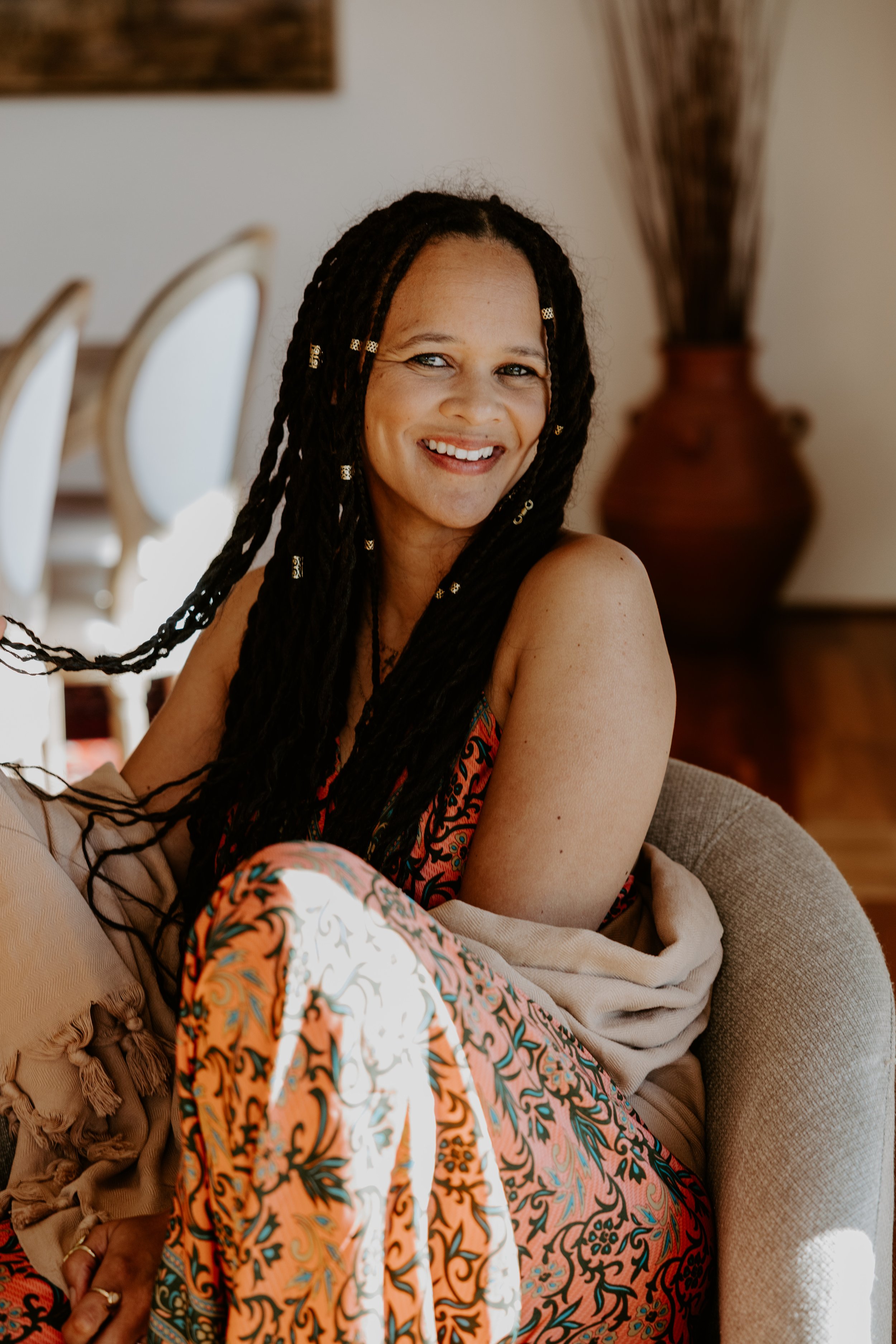 A woman with long, braided hair decorated with gold beads, smiling warmly indoors with a large vase and chairs in the background.