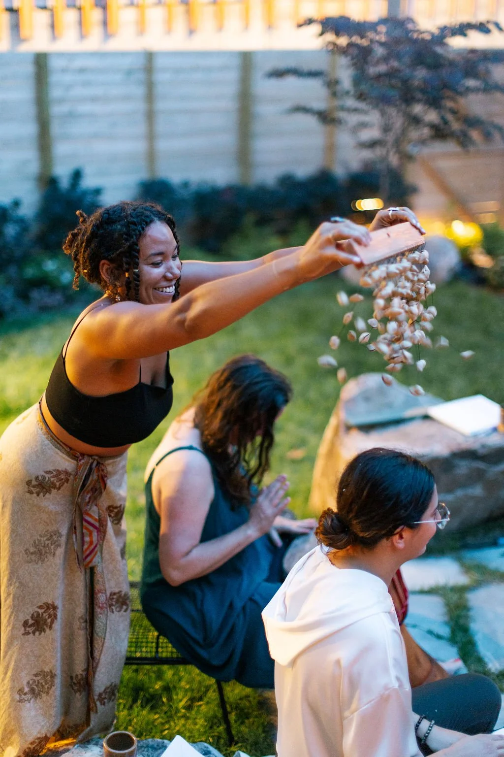 Woman in black top and beige skirt pouring popcorn onto seated women during outdoor gathering in evening.