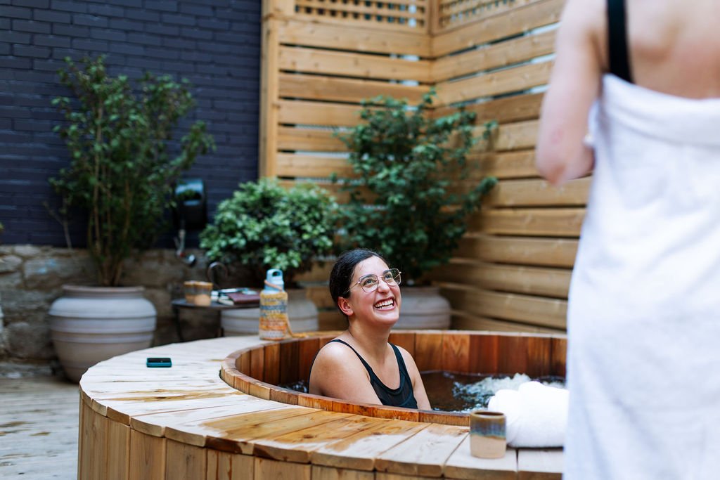 A woman with glasses smiling and sitting in a wooden bathtub outside, with greenery and a wooden fence behind her, while another person stands nearby.