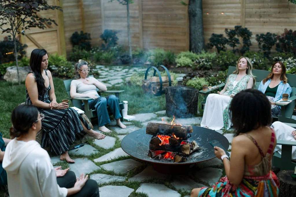 Group of women sitting around a fire pit on a backyard patio, some with eyes closed and some looking at their phones.