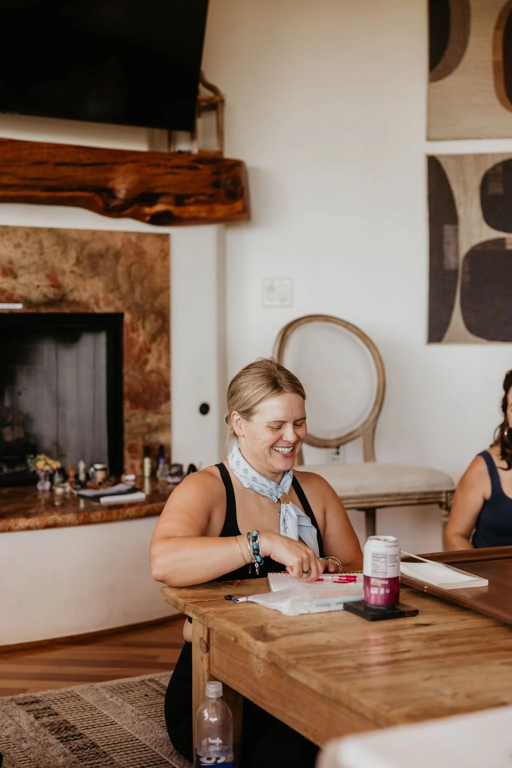 A smiling woman with blonde hair, wearing a black top and a light blue bandana, sitting at a wooden table with a notebook, pens, and a pill bottle, in a cozy living room with a fireplace and decorative wall art.