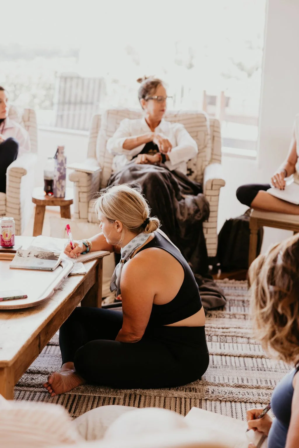 Group of women participating in a meeting or discussion indoors, seated on chairs and a rug, with notes and beverages on a table.