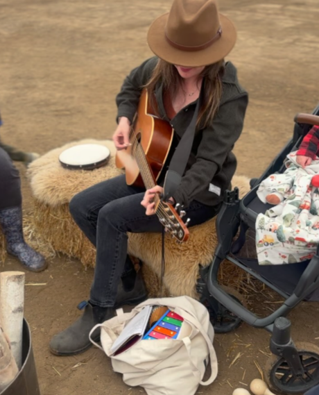 A woman wearing a brown hat and black jacket is sitting on a fluffy stool playing an acoustic guitar. Nearby, a baby is sitting in a stroller wrapped in a blanket, and there is a bag with colorful Rainbow xylophone and sheet music on the ground.