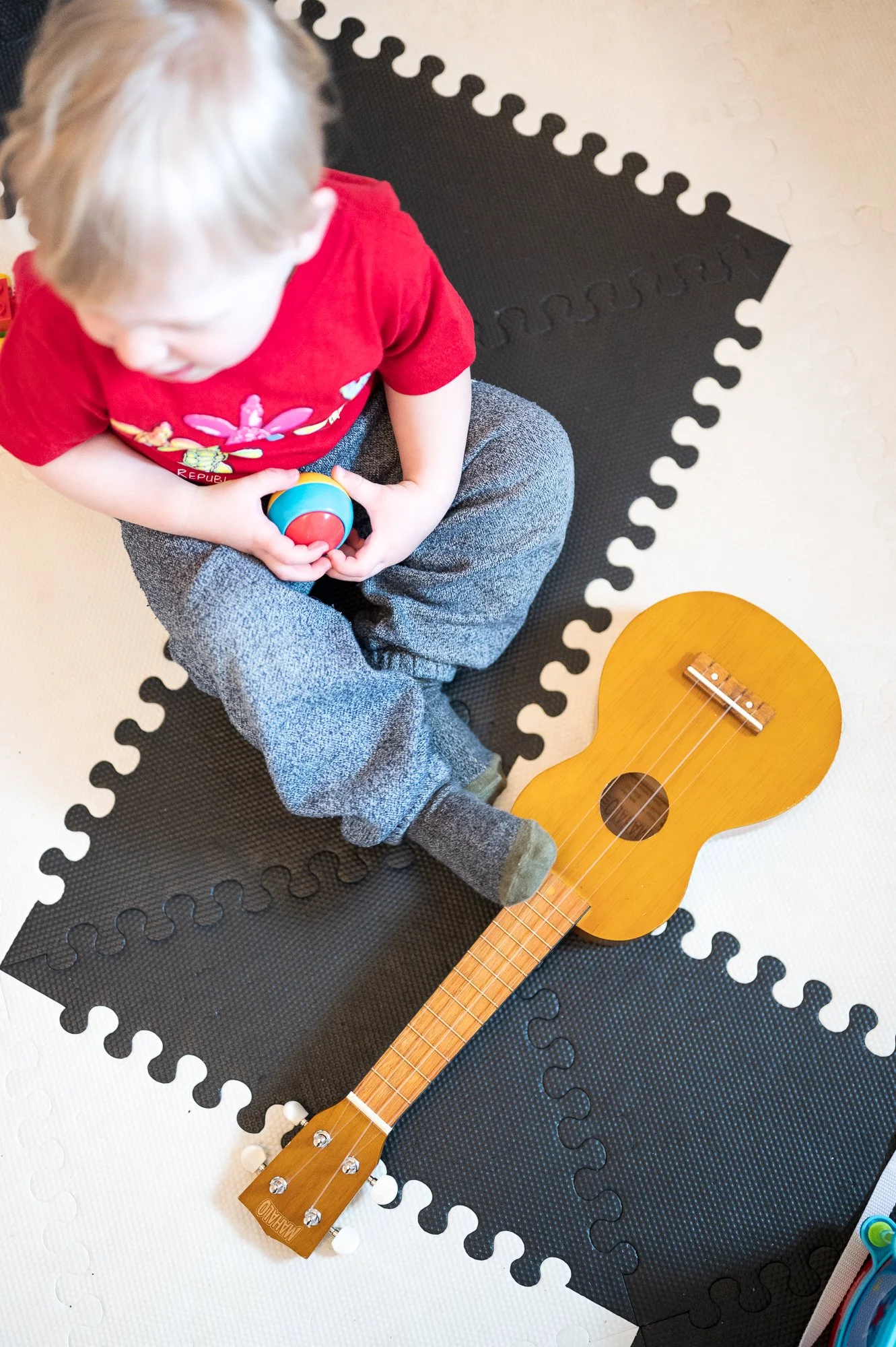 A young child wearing a red shirt and gray pants sitting cross-legged on a black foam mat, holding a colorful toy in his hands. A small wooden ukulele rests nearby on the mat.