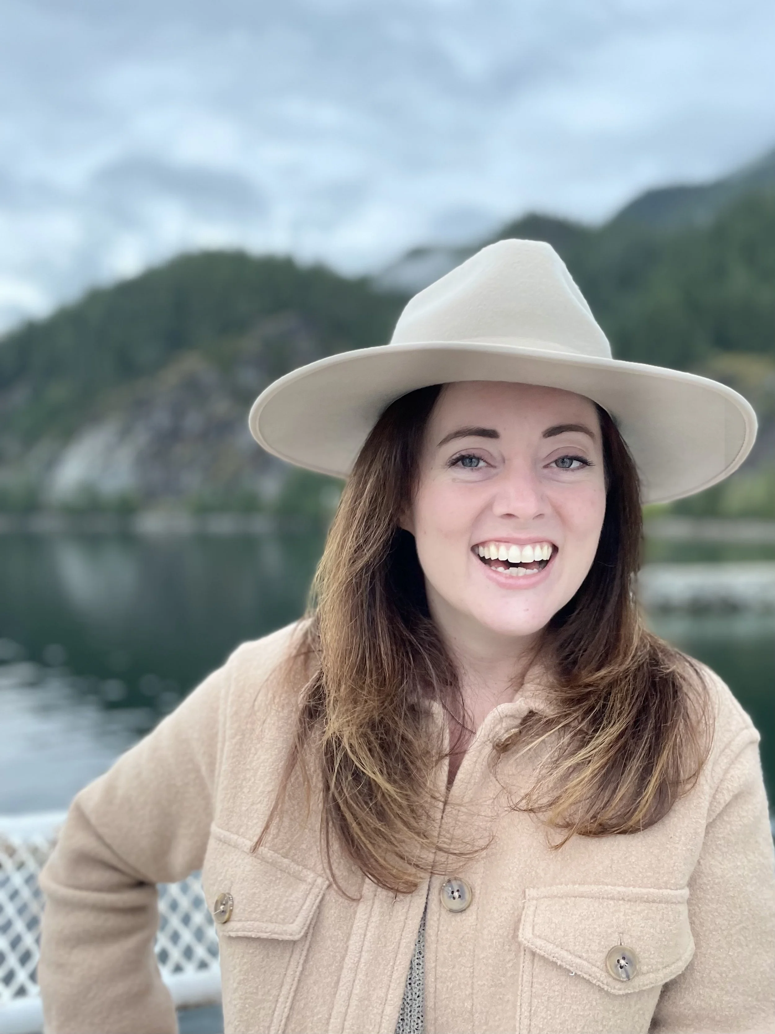 Close-up photo of a smiling woman wearing a soft beige coat and a large white hat, outdoors with water, hills, and cloudy sky in the background.