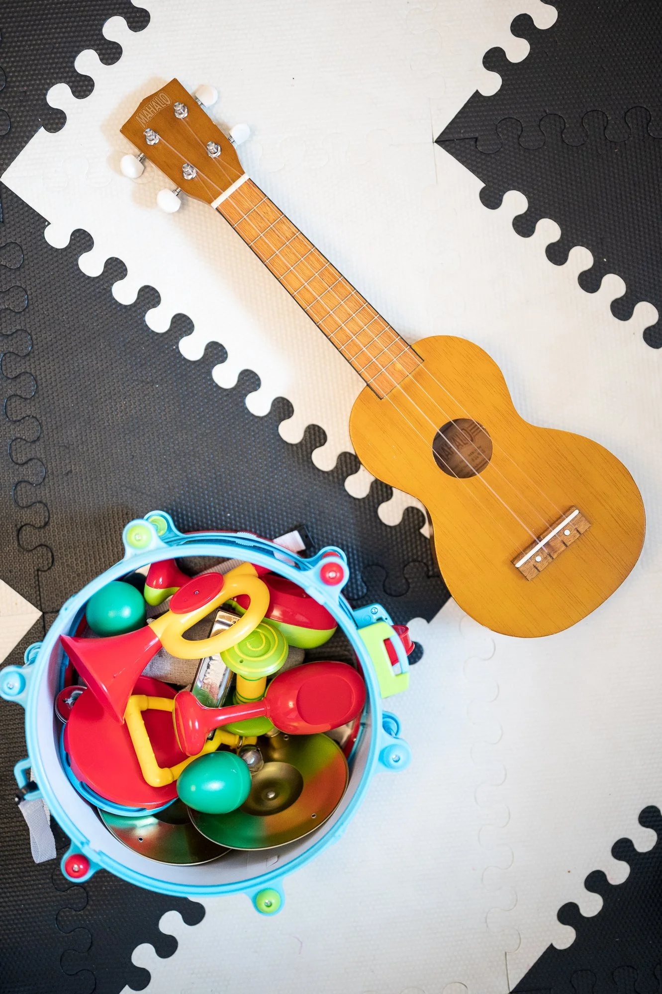 A wooden ukulele resting on a black and white interlocking foam floor tiles next to a colorful plastic bucket filled with various toy musical instruments and accessories.