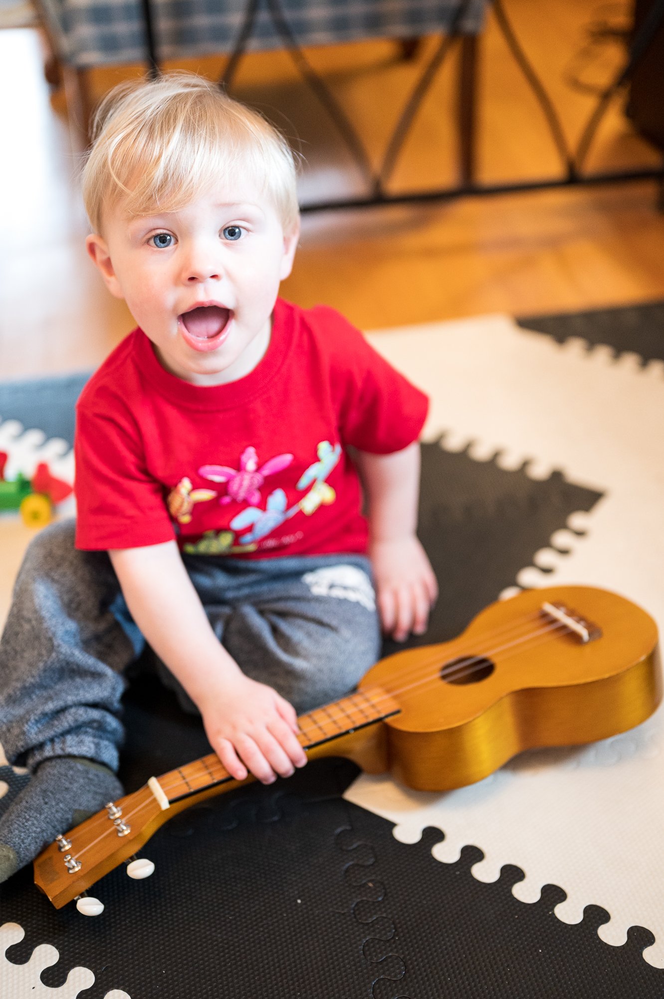 A young boy with blonde hair and blue eyes, wearing a red shirt with colorful butterflies, sitting on a black and white foam puzzle mat. He has an open-mouthed expression and is sitting next to a small wooden guitar placed on the mat.