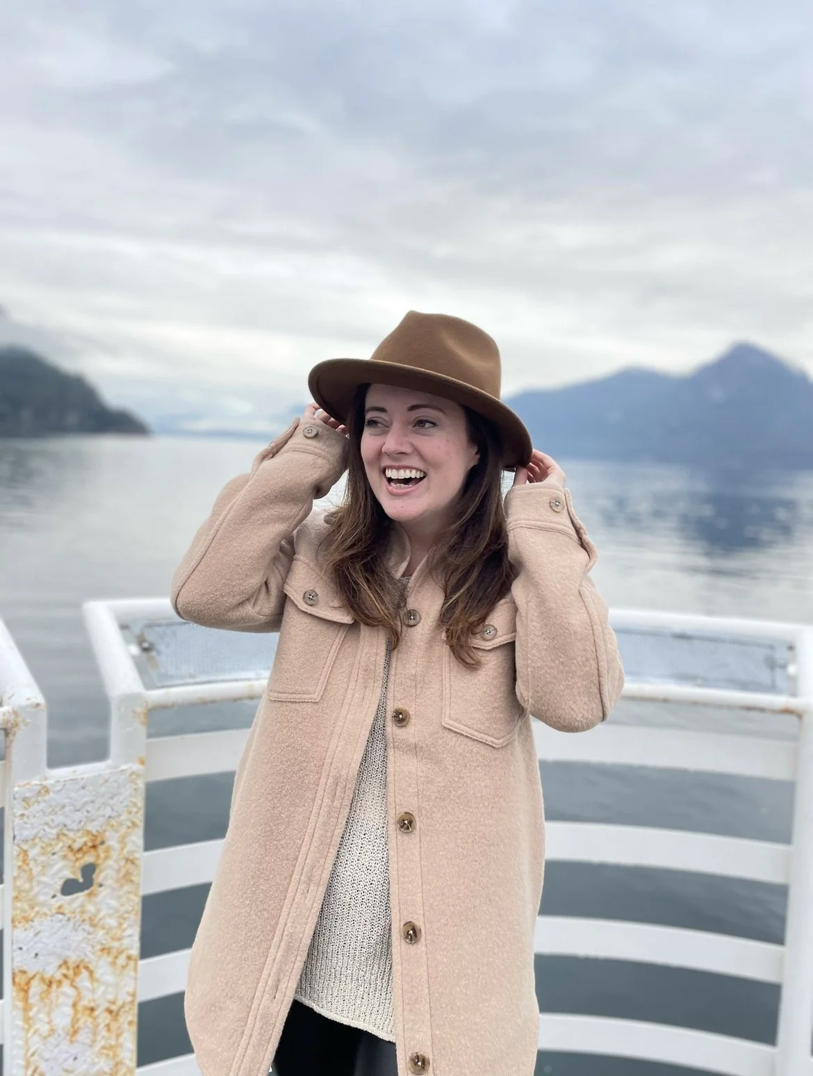 A woman with brown hair wearing a beige coat and a brown hat standing on a boat with water and mountains in the background.