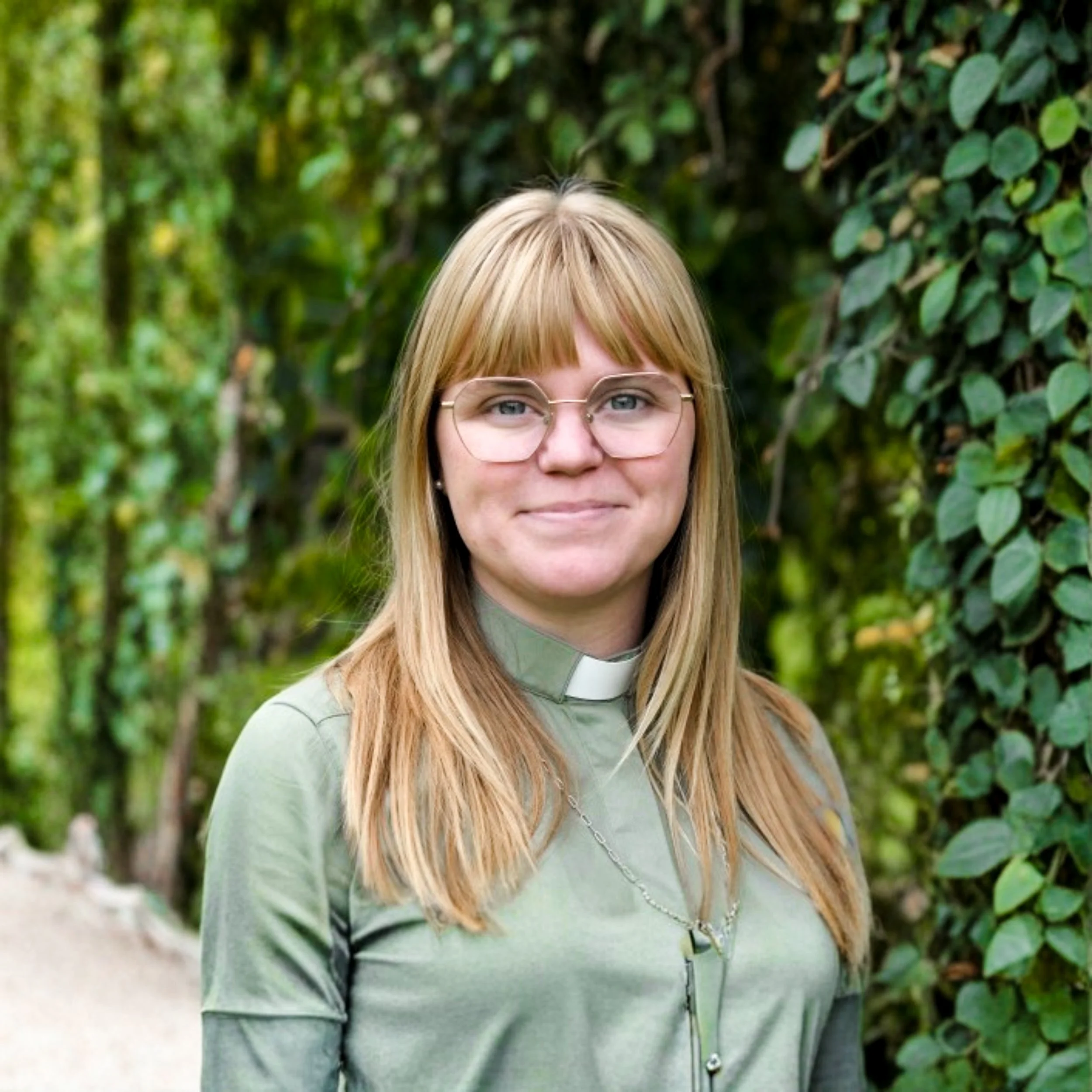 Vilma Bernspång iförd grön diakonskjorta utomhus. I bakgrunden syns lummiga träd och buskar.