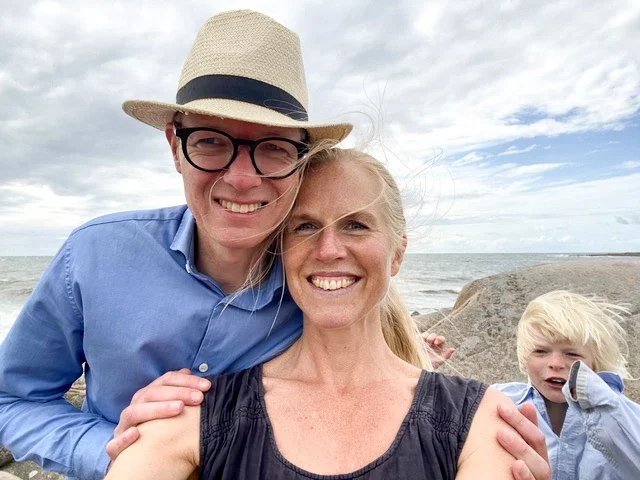 En glad familj på stranden, två vuxna och ett barn, med hav och stenar i bakgrunden.