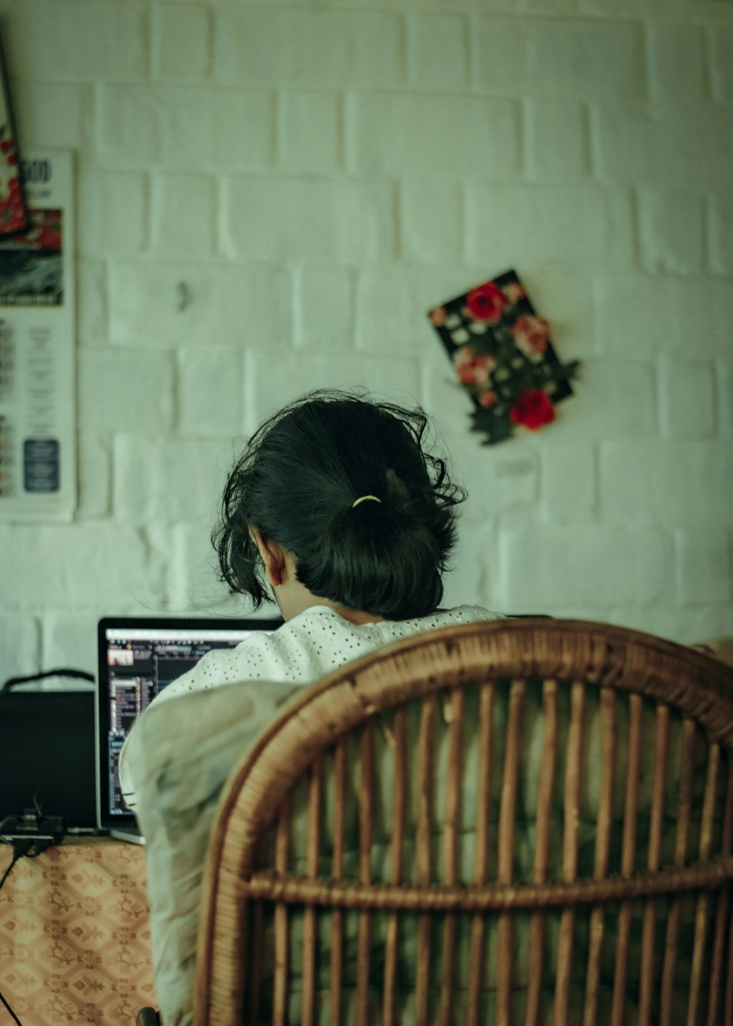 A person with black hair tied back, sitting at a computer desk, using a laptop, viewed from behind. The person is seated in a wicker chair. The background shows a white brick wall with a floral decoration hanging on it, and part of a framed picture or poster is visible on the left side.