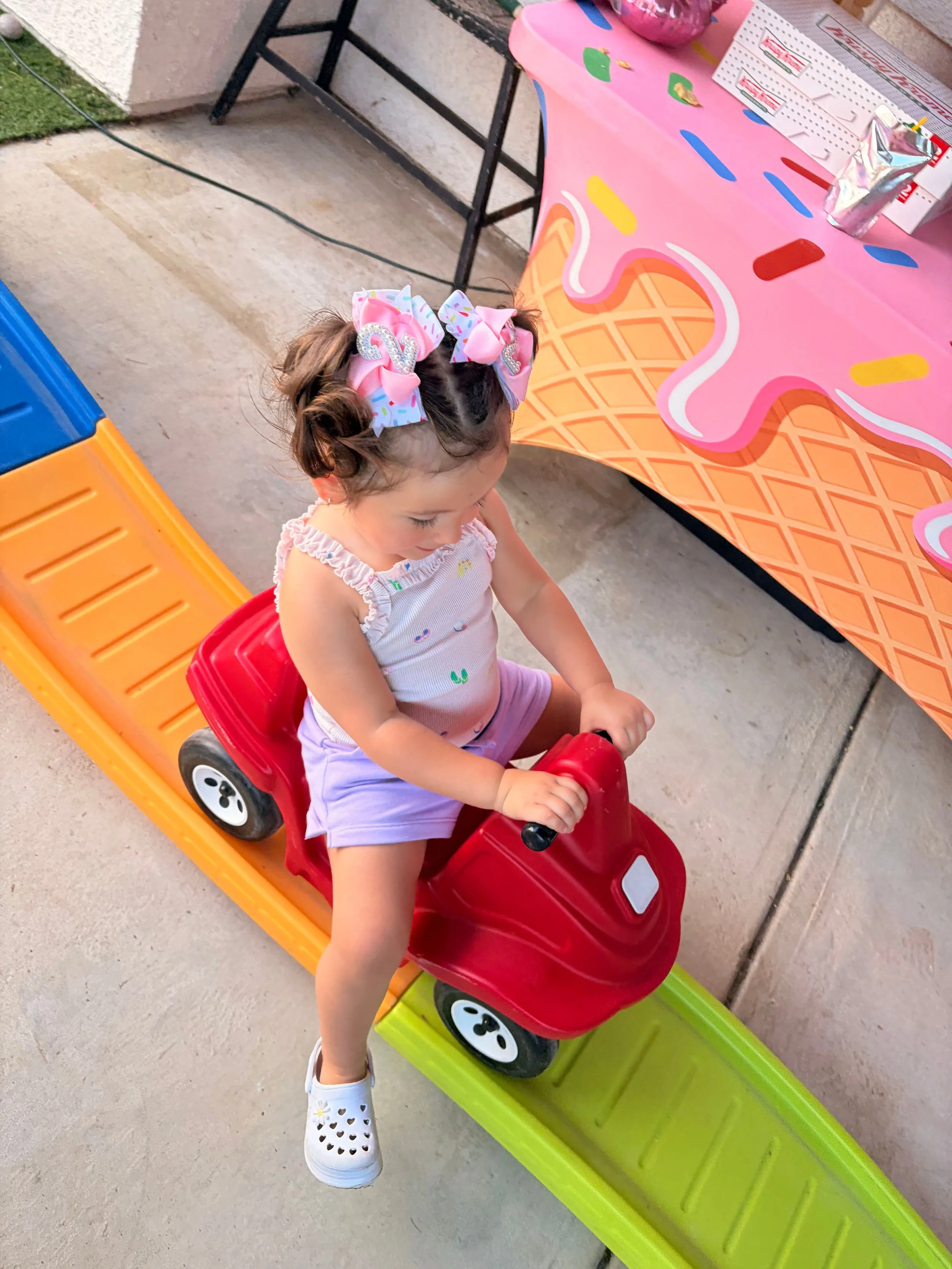 A young girl riding a red and black toy car on a green platform, wearing white heart-shaped sandals, a white tank top with colorful sprinkles, and large pink bows in her hair. She is outdoors near colorful plastic tables and chairs.