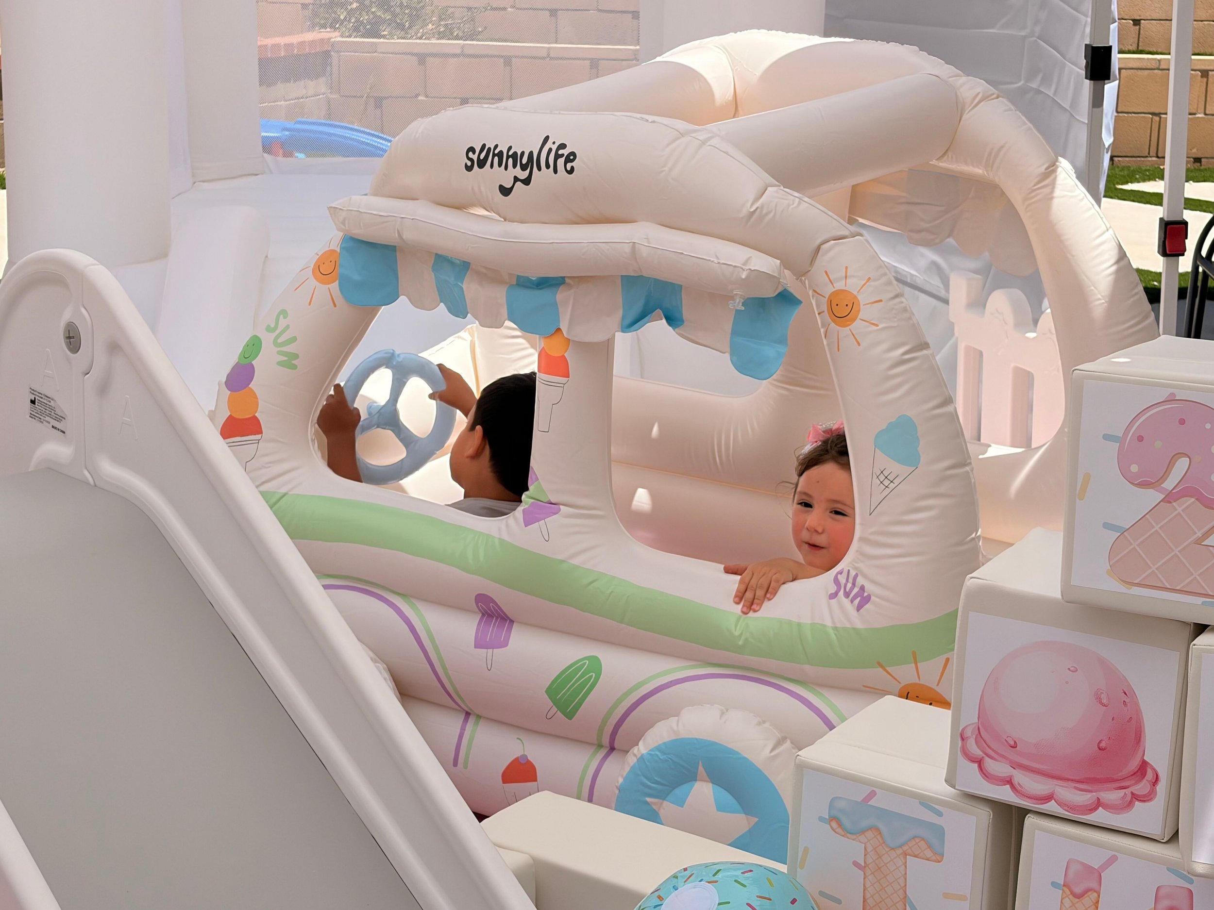 Children playing inside a small inflatable playhouse decorated with ice cream and dessert images, including a smiling sun, and the brand name 'sUnhyLife' visible on the inflatable roof.