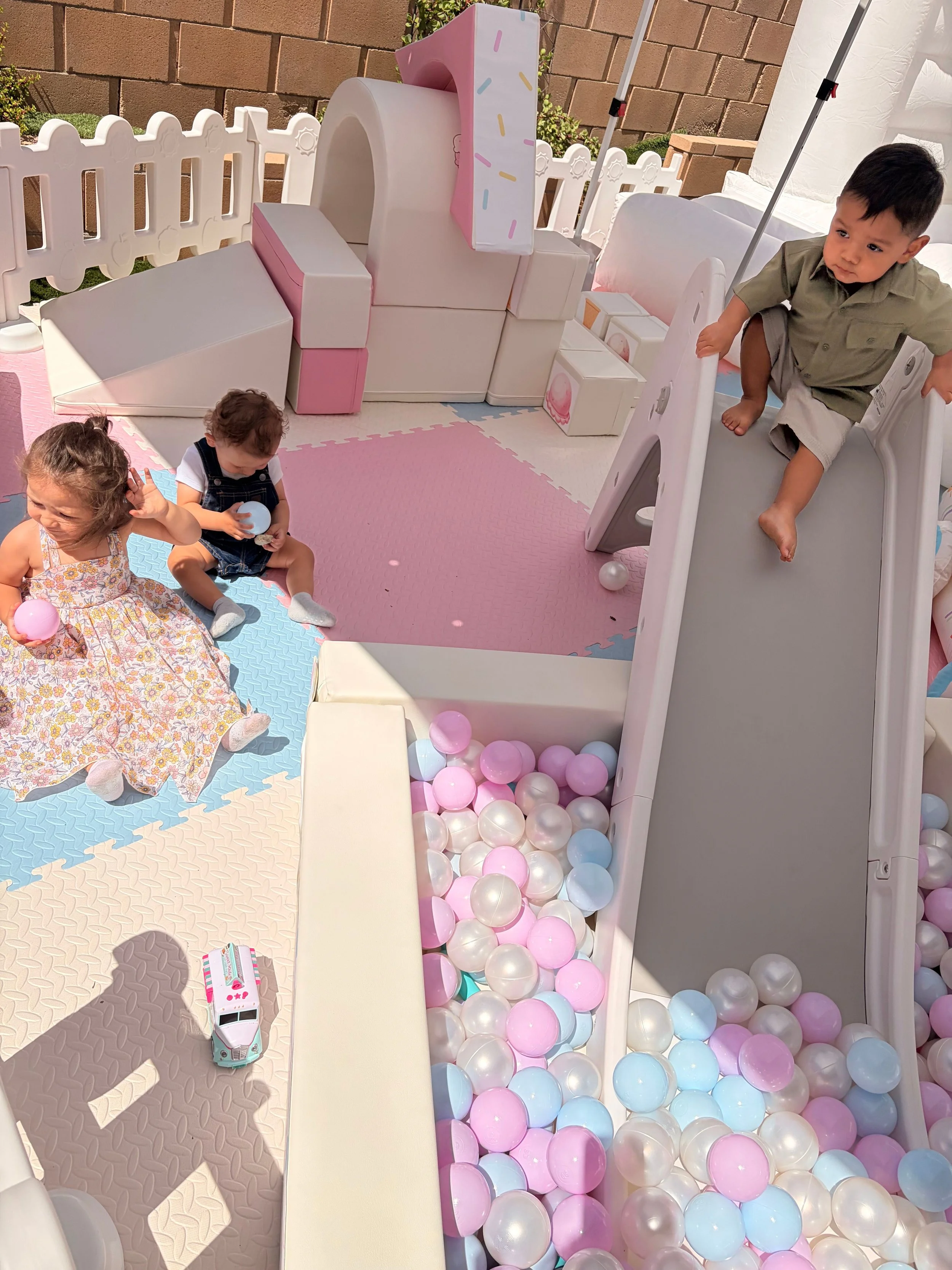 Children playing in a ball pit filled with pink, white, and light blue plastic balls, with a small slide and picnic blanket on the ground.