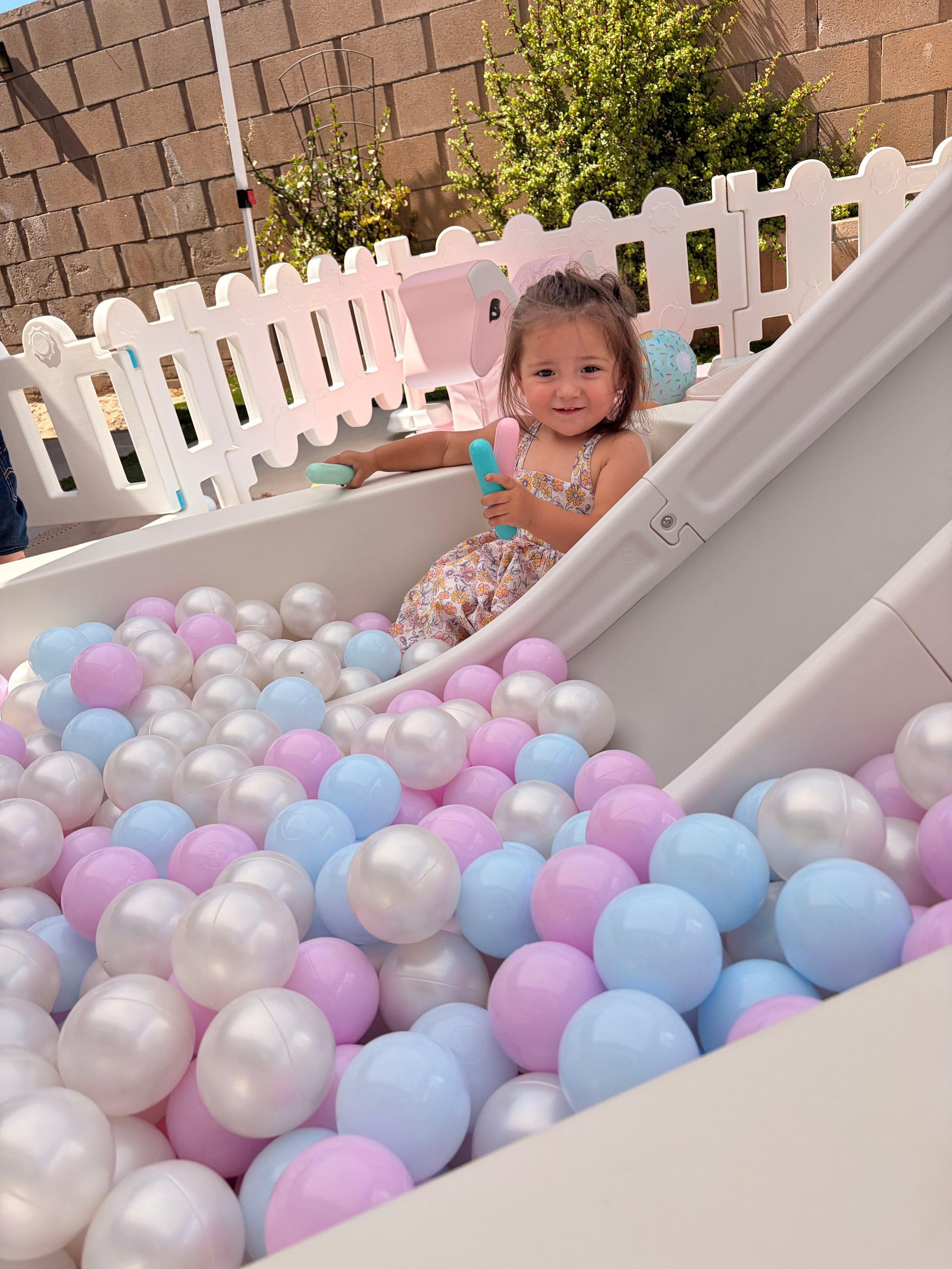 Young girl sitting in a ball pit filled with pastel pink, blue, and white plastic balls, smiling and holding a few balls in her hand, outdoors with a brick wall and green bushes in the background.