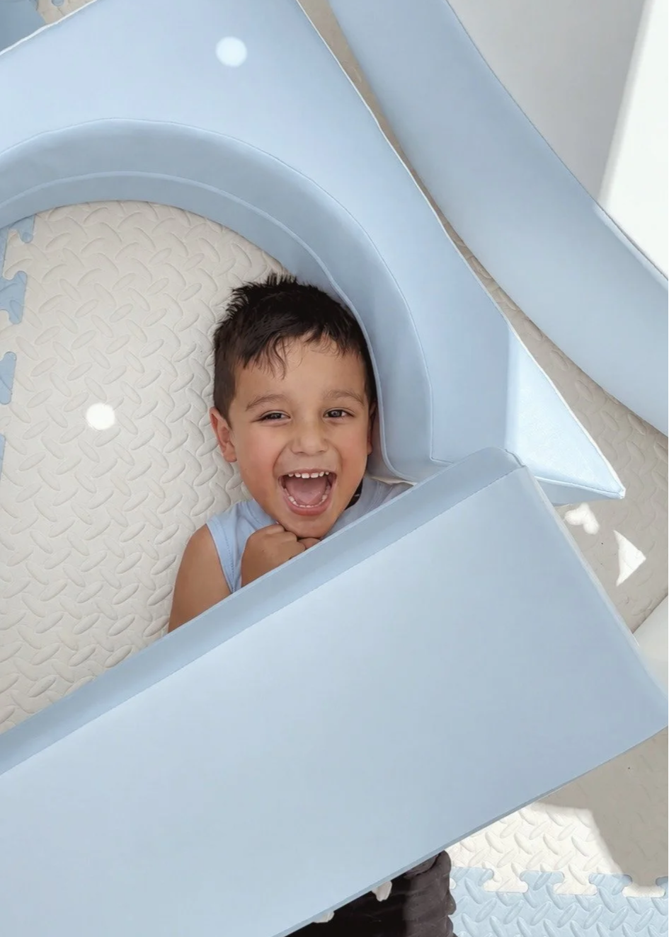 A young boy smiling and laughing while lying in a light blue and white play structure or foam pit with textured flooring.