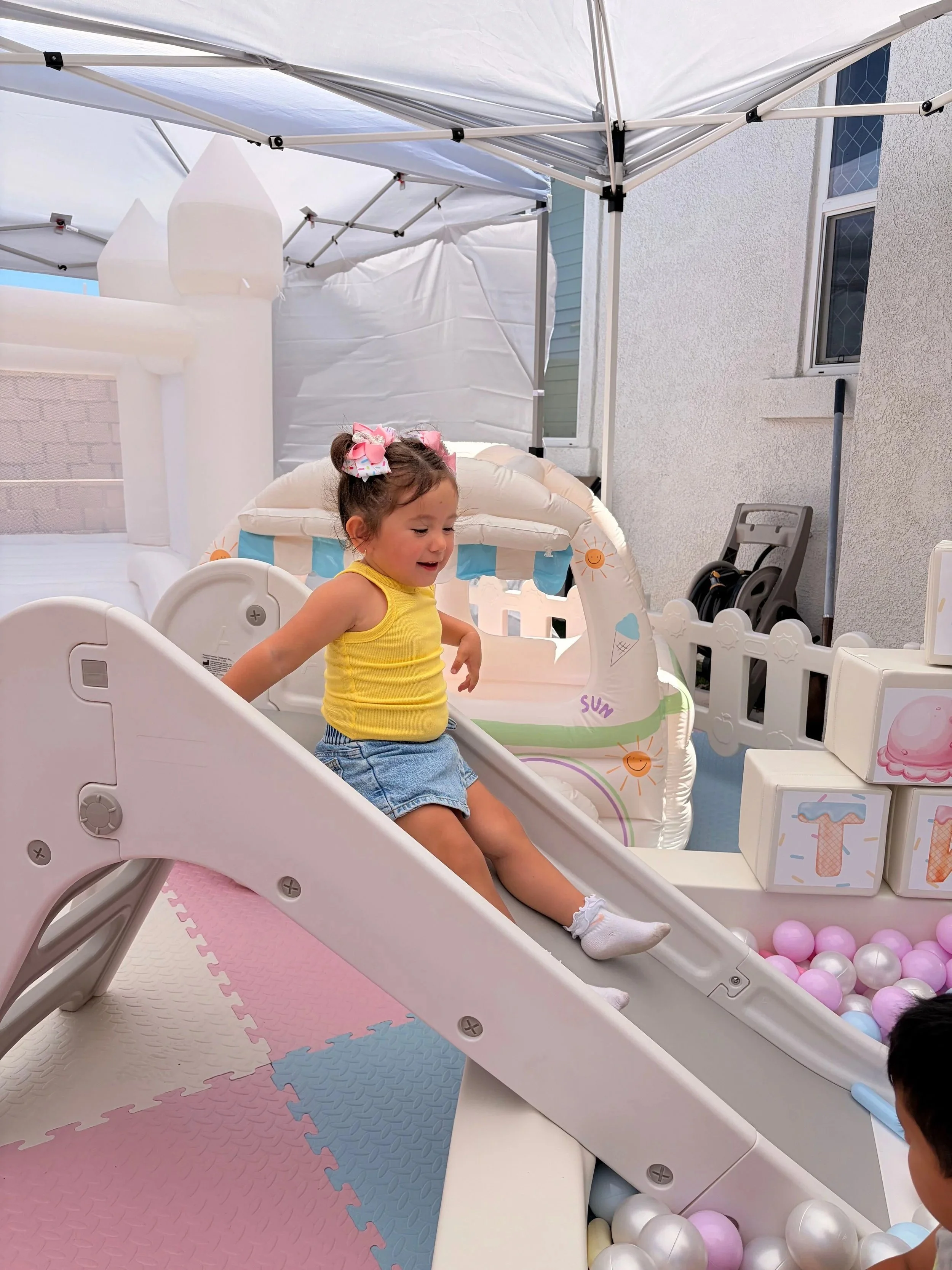 A young girl in a yellow top and denim shorts playing on a small slide in an outdoor play area, surrounded by a colorful foam mat and a ball pit with pink, white, and purple balls, under a white canopy.
